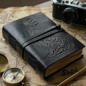 May include: Black leather journal with embossed wolf and moon designs. The journal has a leather strap closure and aged paper pages. A vintage camera, compass, and pen are also visible.