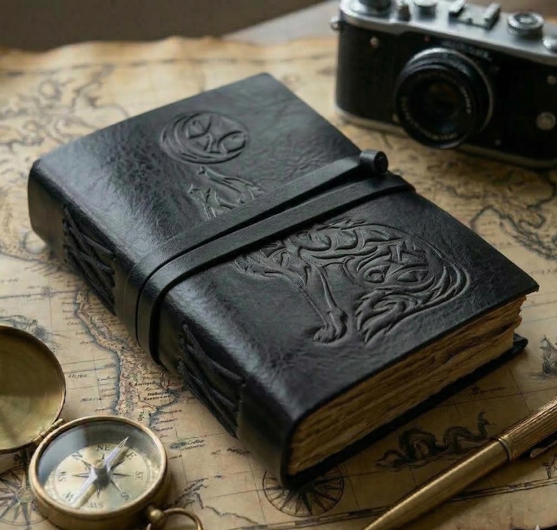 May include: Black leather journal with embossed wolf and moon designs. The journal has a leather strap closure and aged paper pages. A vintage camera, compass, and pen are also visible.