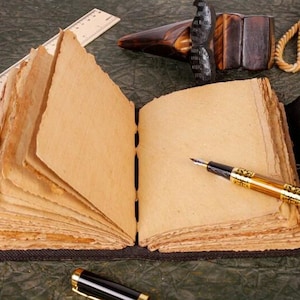 May include: A brown leather-bound journal with blank pages, a gold-toned fountain pen, and a wooden carving on a green surface.