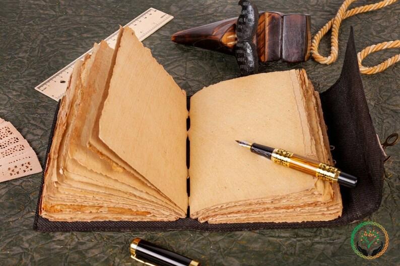 May include: A brown leather-bound journal with blank pages, a gold-toned fountain pen, and a wooden carving on a green surface.