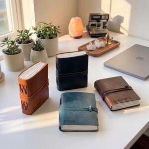 May include: A collection of leather-bound journals in various colors, including brown, black, and teal, each with a name embossed on the cover. The journals are closed with a leather strap. The journals are displayed on a white surface.