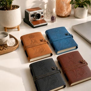 May include: Four leather journals in brown, blue, black, and dark brown, each with a debossed wolf design and a leather strap closure. A vintage camera and plants are in the background.