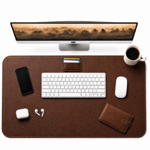 Könnte beinhalten: Eine braune Schreibtischunterlage mit Tastatur, Maus, Telefon, Geldbörse und einer Tasse Kaffee. Ein Computermonitor zeigt ein Landschaftsbild. Ein kleiner Kartenhalter aus Leder ist an der Unterlage befestigt. Weiße kabellose Ohrhörer und ein Ladecase sind ebenfalls vorhanden.