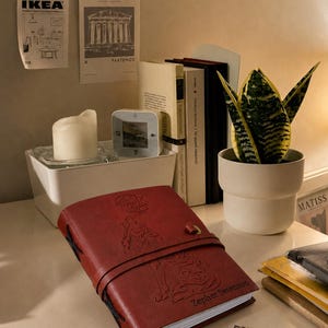 May include: A red leather journal with an embossed wolf design and the name "Zepher Swenson" is displayed on a desk. A small white candle, a potted snake plant, and several books are also visible.