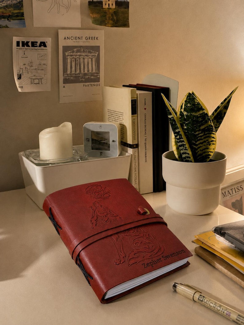May include: A red leather journal with an embossed wolf design and the name "Zepher Swenson" is displayed on a desk. A small white candle, a potted snake plant, and several books are also visible.