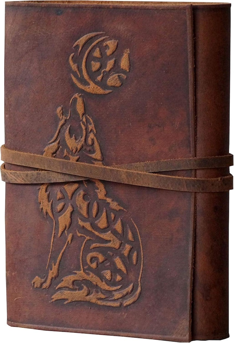 May include: A brown leather journal with a wolf and moon design. The cover features an embossed wolf howling at the moon, with a leather strap closure. The journal has a rustic, vintage aesthetic.