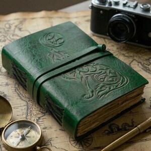 May include: A green leather journal with a wolf and moon design embossed on the cover. The journal is closed with a leather strap. A vintage camera and compass are in the background.