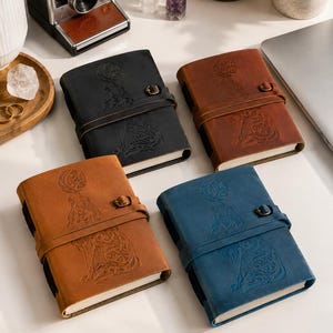 May include: Four leather journals in black, brown, tan, and blue. Each journal features an embossed design of a figure with a moon and animal, secured with a leather strap and metal clasp. The journals are on a white surface.