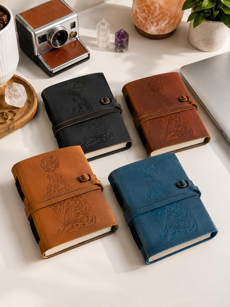 May include: Four leather journals in black, brown, tan, and blue. Each journal features an embossed design of a figure with a moon and animal, secured with a leather strap and metal clasp. The journals are on a white surface.