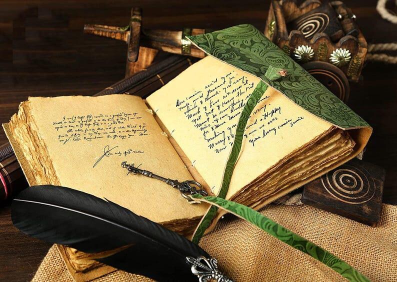 Puede incluir: Un diario de estilo vintage con una cubierta de cuero verde y una pluma de ave. El diario tiene un aspecto desgastado y antiguo y est&aacute; abierto para revelar p&aacute;ginas en blanco.