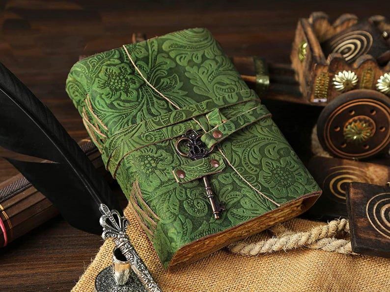 Puede incluir: Un diario de cuero verde con un dise&ntilde;o floral en relieve y un cierre en forma de llave. El diario est&aacute; cerrado y se encuentra sobre una superficie marr&oacute;n.