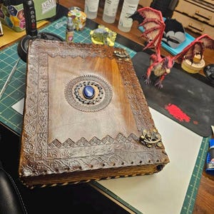 May include: A large, brown leather-bound book with intricate embossed designs and a central blue gemstone. The book features decorative metal clasps and a detailed border. A red dragon figurine is visible nearby.