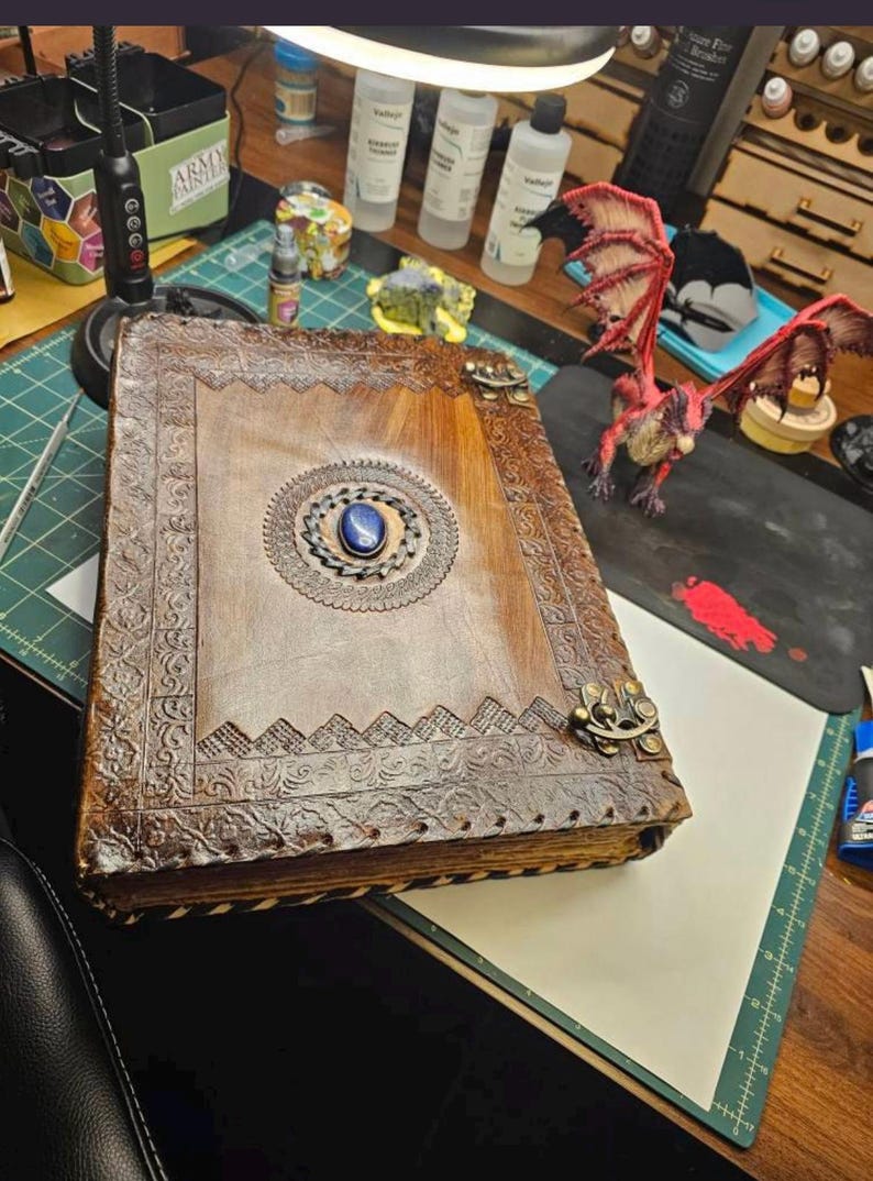 May include: A large, brown leather-bound book with intricate embossed designs and a central blue gemstone. The book features decorative metal clasps and a detailed border. A red dragon figurine is visible nearby.