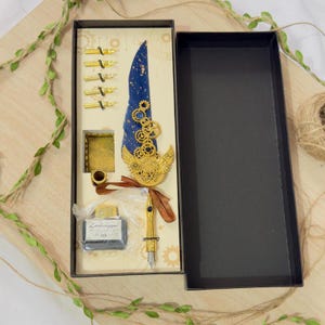 Puede incluir: Un juego de pluma de caligraf&iacute;a de estilo steampunk azul y dorado en una caja. La pluma tiene una pluma azul con engranajes dorados y una punta dorada. El juego incluye un peque&ntilde;o frasco de tinta, un portaplumas y varias plumas.