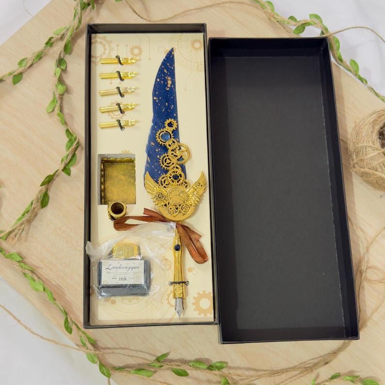 Puede incluir: Un juego de pluma de caligraf&iacute;a de estilo steampunk azul y dorado en una caja. La pluma tiene una pluma azul con engranajes dorados y una punta dorada. El juego incluye un peque&ntilde;o frasco de tinta, un portaplumas y varias plumas.