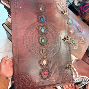 May include: A large, brown leather journal with intricate embossed designs and colorful gemstone accents. The journal features a series of decorative circles and a central pattern, secured by metal clasps. The edges are stitched with a thick thread.