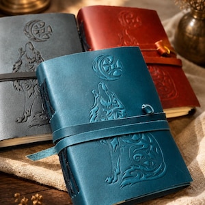 May include: Leather-bound journals in shades of blue, gray, and red. Each journal features an embossed design of a howling wolf under a crescent moon. The journals have a strap closure.