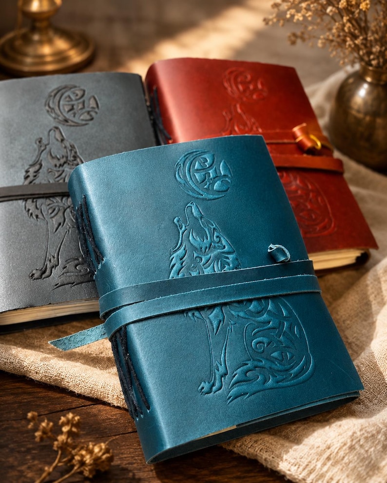 May include: Leather-bound journals in shades of blue, gray, and red. Each journal features an embossed design of a howling wolf under a crescent moon. The journals have a strap closure.