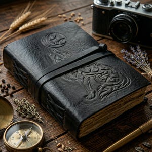 May include: A black leather journal with an embossed wolf design and a strap closure. The journal is on a wooden surface with a vintage camera, compass, and pen.