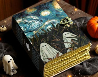 May include: A decorative journal with a Halloween theme. The cover features a starry night sky and two ghost illustrations. The journal has gold-colored edges and a metal clasp. The journal is on a wooden surface with small pumpkins.