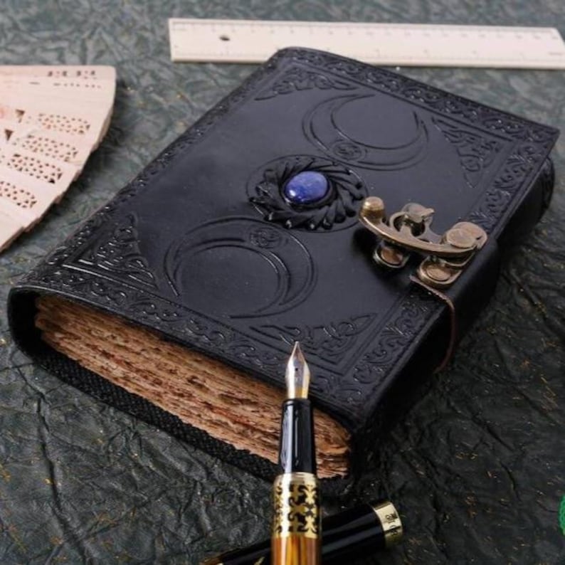 May include: A black leather journal with a decorative metal clasp and a blue gemstone. The journal has a detailed embossed design of three crescent moons. The journal is open to reveal blank pages.