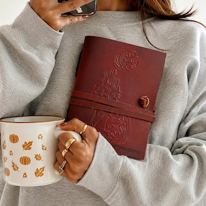 May include: A maroon leather journal with a wolf and moon design, secured by a leather strap and a brass clasp. A white mug with orange pumpkin and leaf designs is held in the other hand.