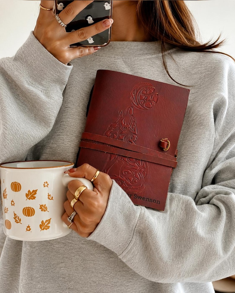 May include: A maroon leather journal with a wolf and moon design, secured by a leather strap and a brass clasp. A white mug with orange pumpkin and leaf designs is held in the other hand.