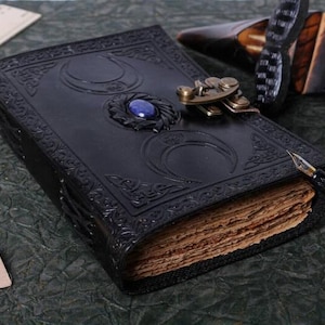 May include: A black leather journal with a decorative metal clasp and a blue gemstone. The journal has a triple moon design embossed on the cover and is closed with a metal clasp. The journal is lying on a green surface with a wooden pen and a ruler.
