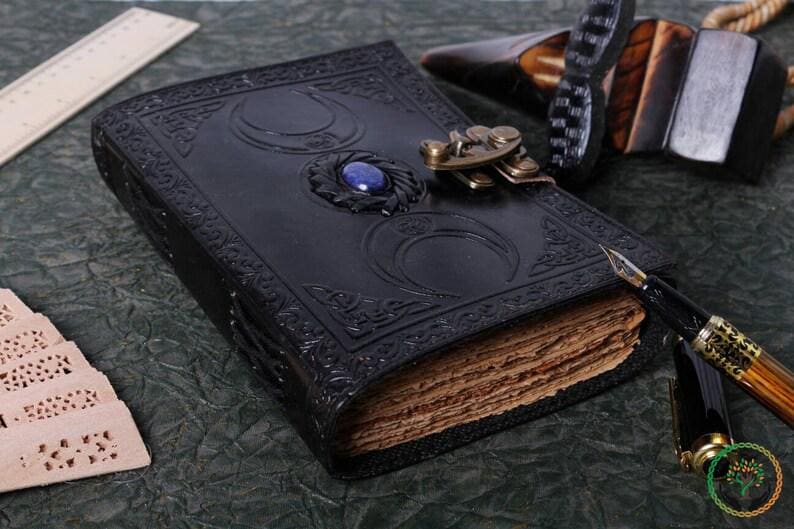 May include: A black leather journal with a decorative metal clasp and a blue gemstone. The journal has a triple moon design embossed on the cover and is closed with a metal clasp. The journal is lying on a green surface with a wooden pen and a ruler.