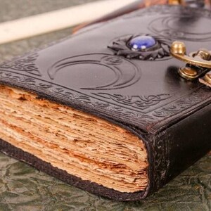 May include: A brown leather journal with a decorative moon design and a blue gemstone. The journal has a clasp closure and is closed.