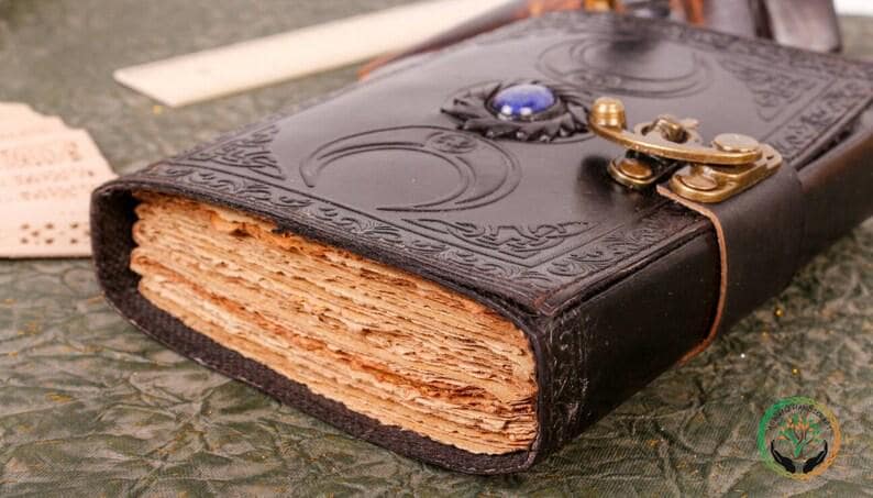 May include: A brown leather journal with a decorative moon design and a blue gemstone. The journal has a clasp closure and is closed.