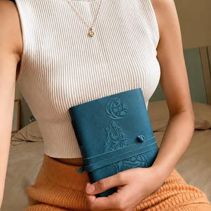 May include: Teal-colored journal with an embossed wolf and moon design. The journal is held by a person wearing a white ribbed tank top and a gold necklace. The person is also wearing an orange skirt.