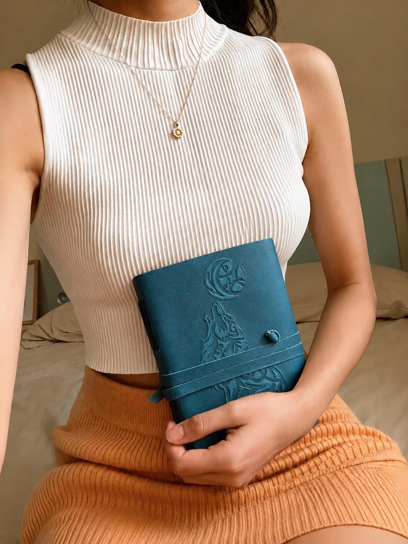 May include: Teal-colored journal with an embossed wolf and moon design. The journal is held by a person wearing a white ribbed tank top and a gold necklace. The person is also wearing an orange skirt.