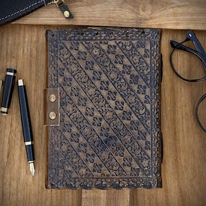May include: A brown, intricately carved journal with a leather strap closure. A black fountain pen and a small black leather wallet are next to the journal on a wooden surface. Black eyeglasses are also visible.