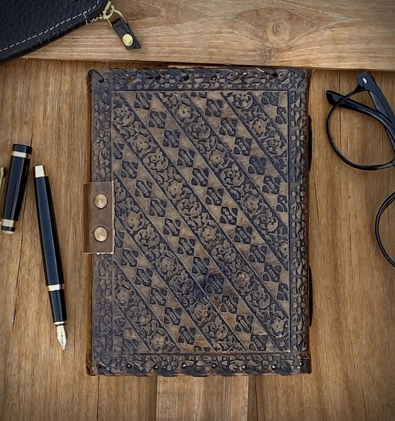 May include: A brown, intricately carved journal with a leather strap closure. A black fountain pen and a small black leather wallet are next to the journal on a wooden surface. Black eyeglasses are also visible.