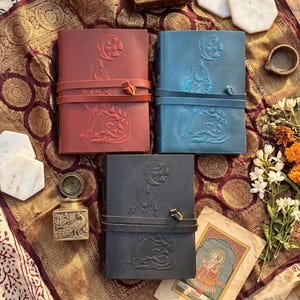 May include: Three leather journals in red, blue, and black, each with a wolf howling at the moon embossed design. The journals have a wrap-around leather strap closure. The image also includes a small decorative box, marble coasters, and a vintage painting.
