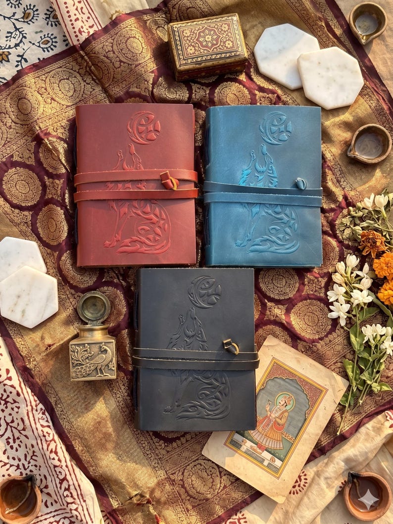 May include: Three leather journals in red, blue, and black, each with a wolf howling at the moon embossed design. The journals have a wrap-around leather strap closure. The image also includes a small decorative box, marble coasters, and a vintage painting.