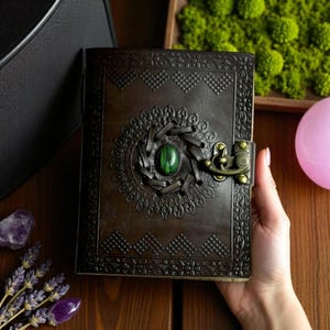 May include: A dark brown leather journal with an embossed floral design and a green stone centerpiece. The journal has a brass clasp and is held by a hand. The journal is a unique and decorative item.