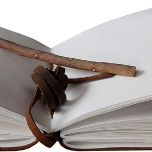 May include: An open, brown leather-bound journal with blank white pages. A small, natural wooden stick rests across the pages. A brown leather strap with a knot closure is attached to the journal.