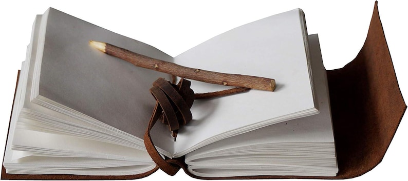 May include: An open, brown leather-bound journal with blank white pages. A small, natural wooden stick rests across the pages. A brown leather strap with a knot closure is attached to the journal.
