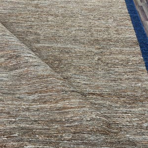 Puede incluir: Una alfombra rectangular con una textura natural tejida en tonos beige y marrón. La alfombra tiene un borde azul oscuro, y una parte de la alfombra está enrollada, revelando su parte inferior. La alfombra parece estar hecha de fibras naturales.