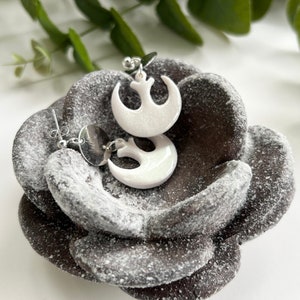 May include: A pair of white Rebel Alliance symbol earrings with silver posts. The earrings are displayed on a dark brown, textured surface.