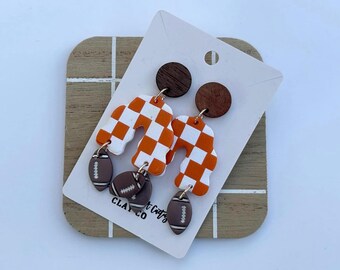 May include: Orange and white checkered arch-shaped earrings with brown football charms. The earrings are hanging from brown wooden circles.