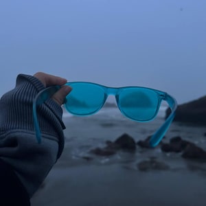 Op de afbeelding: Doorschijnende felblauwe zonnebril met een klassieke montuurvorm. De monturen hebben een gladde, half-transparante afwerking waardoor licht door de glazen valt, wat een levendige cyaan tint creëert bij natuurlijk buitenlicht.