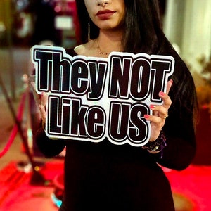 May include: A person holding a sign that says "They Not Like Us" in black and white letters. The person is wearing a black dress and has long dark hair.