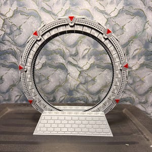 May include: A detailed, gray model of a Stargate, featuring a circular design with intricate patterns and red triangular accents. The model sits on a rectangular base with a brick-like pattern, showcasing a sci-fi theme.