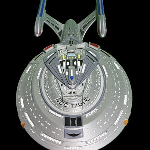 May include: A model of the USS Enterprise starship, featuring a gray saucer section with numerous white lights and the text "NCC-1701-E." The ship has two warp nacelles with blue and red accents, and a gray secondary hull. The model is set against a black background.