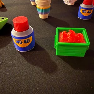 May include: A collection of miniature novelty items, including a coffee cup with a colorful sleeve, a small green container filled with red material, and a blue WD-40 bottle with red cap. Other items include a skull and a strawberry.