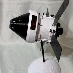 May include: A 3D-printed model of a spacecraft, featuring a black and white color scheme with solar panels. The model is mounted on a white, textured base, and the text "NASA" is visible on the side.