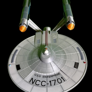 Puede incluir: Un modelo plateado de la nave espacial U.S.S. Enterprise NCC-1701, con un casco principal circular y dos góndolas de curvatura. El modelo tiene líneas de panel detalladas y el nombre de la nave está impreso en el casco. Las góndolas tienen detalles en naranja y azul.